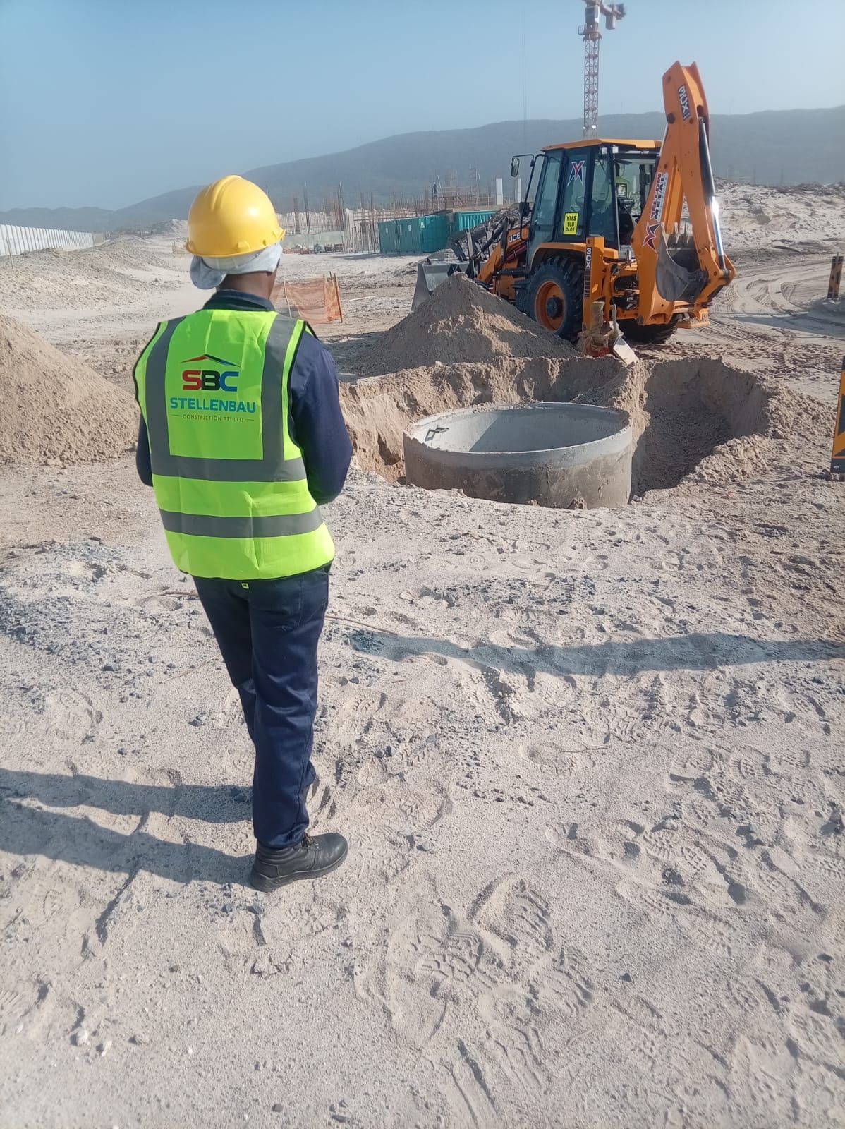 SBC site supervisor in a branded hi-vis vest and hard hat overseeing a precast manhole installation